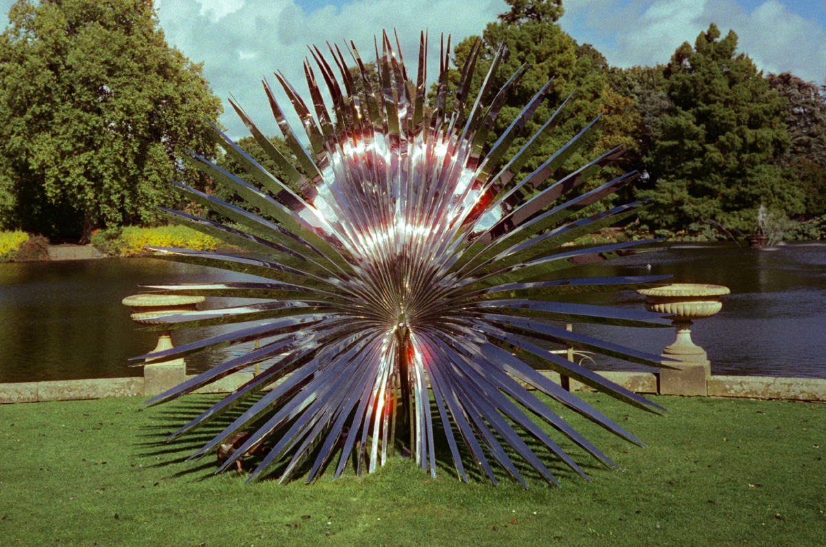 A mirrored sculpture with sharp, radiating metal blades reflecting sunlight, set against a pond and green trees.