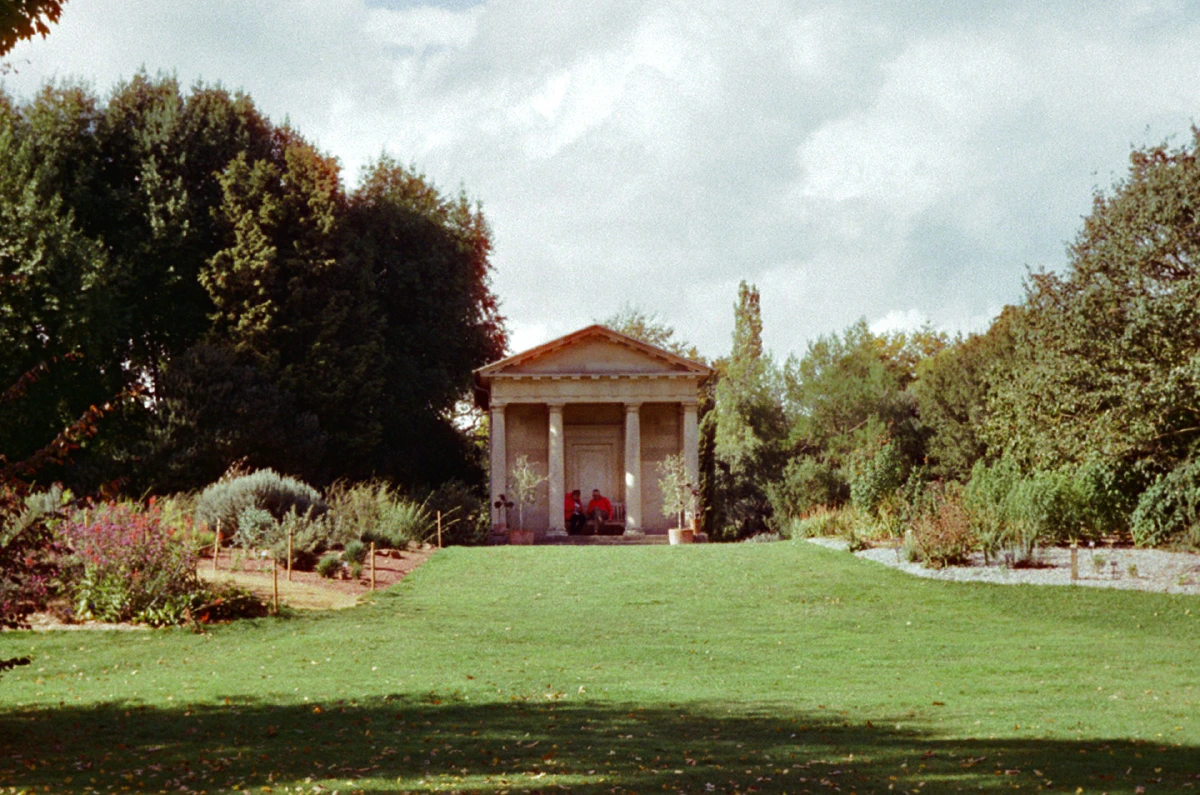 A small classical-style building with columns sits at the top of a grassy hill, two people resting in its shade.