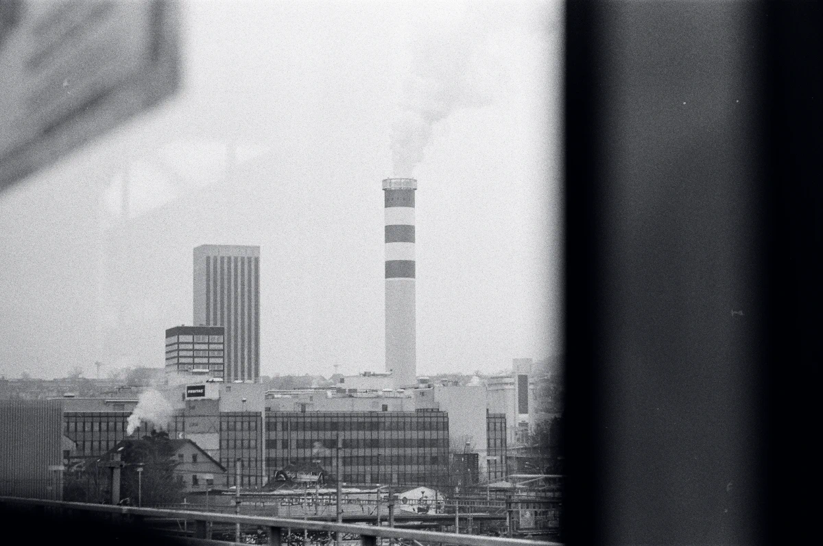 A black and white photograph of an industrial cityscape, featuring a tall smokestack with stripes emitting smoke, surrounded by office buildings and warehouses, viewed through the window of a moving tram.