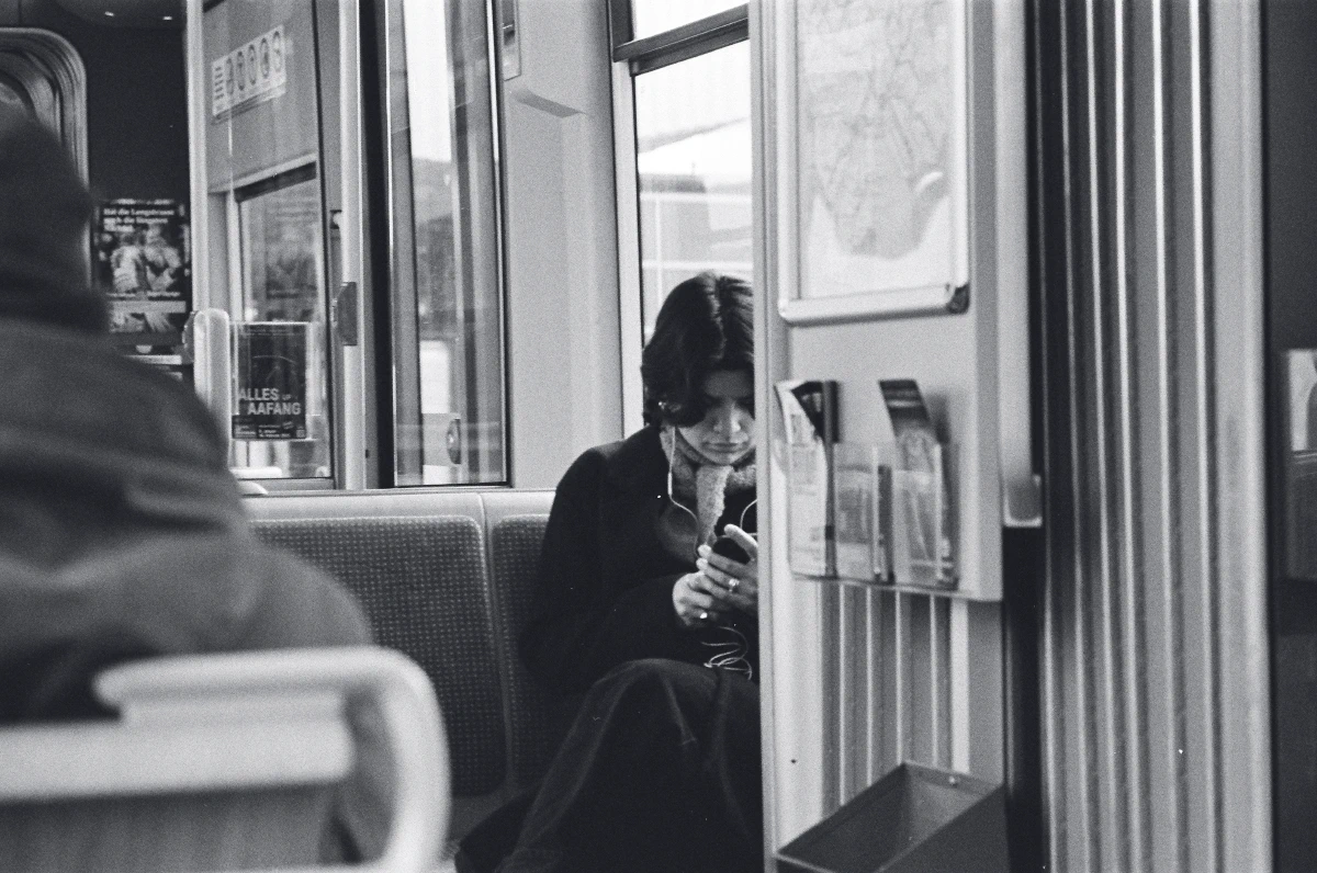 A black and white photograph of a woman sitting inside a tram, looking at her phone, with her headphones untangled on her lap.