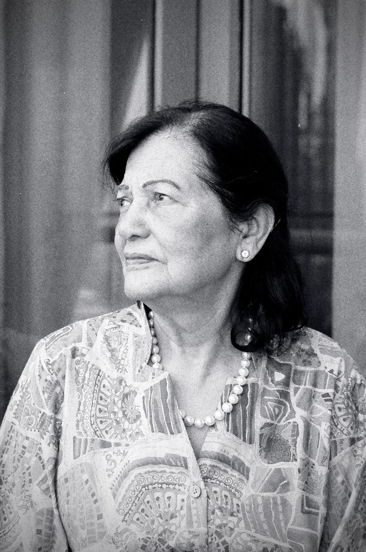 1/2: A black and white portrait of my grandmother, dressed in a patterned blouse with a pearl necklace, gazing thoughtfully to the side, with a serene expression.