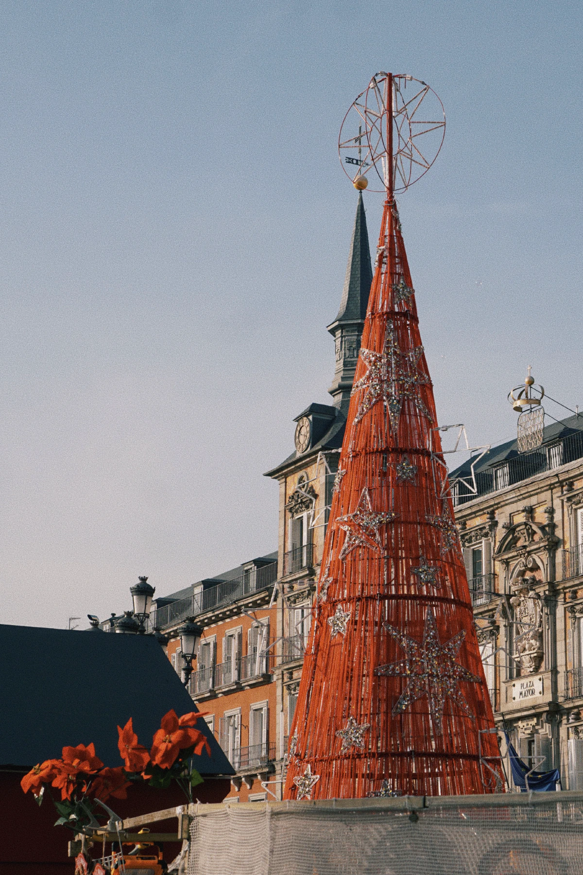 A large red wire-frame Christmas tree adorned with star decorations in a festive plaza, with historic buildings and poinsettias in the foreground.