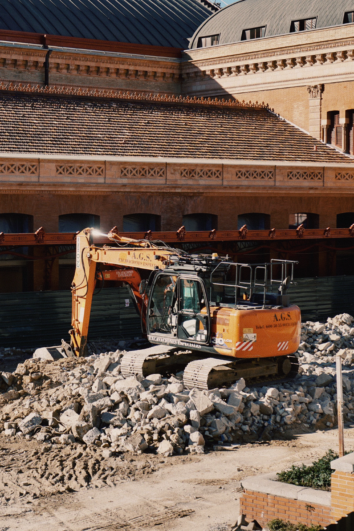 An orange excavator parked atop a pile of rubble at a construction site in front of a historic brick building with arched windows.