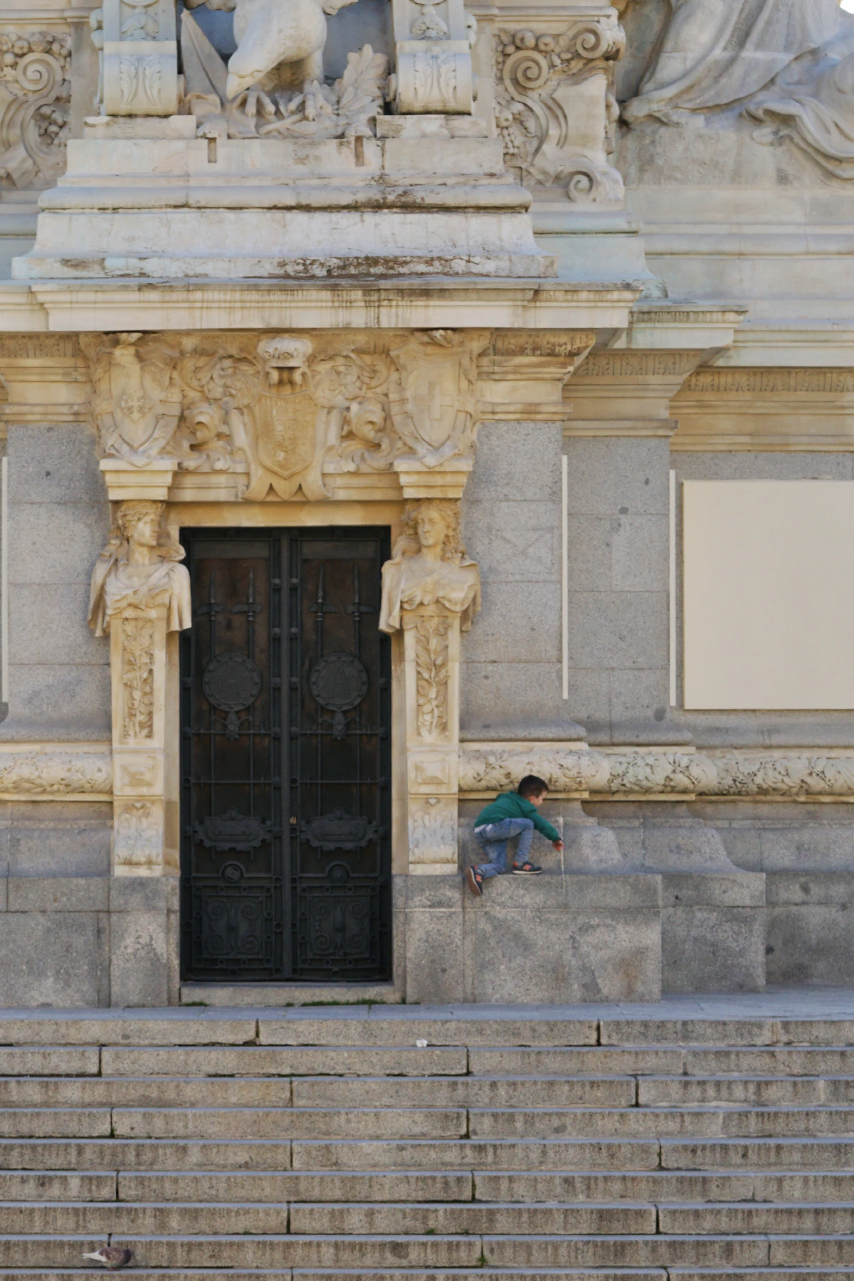 A young child climbing a stone ledge in front of a historic building adorned with classical sculptures and a large ornate door.