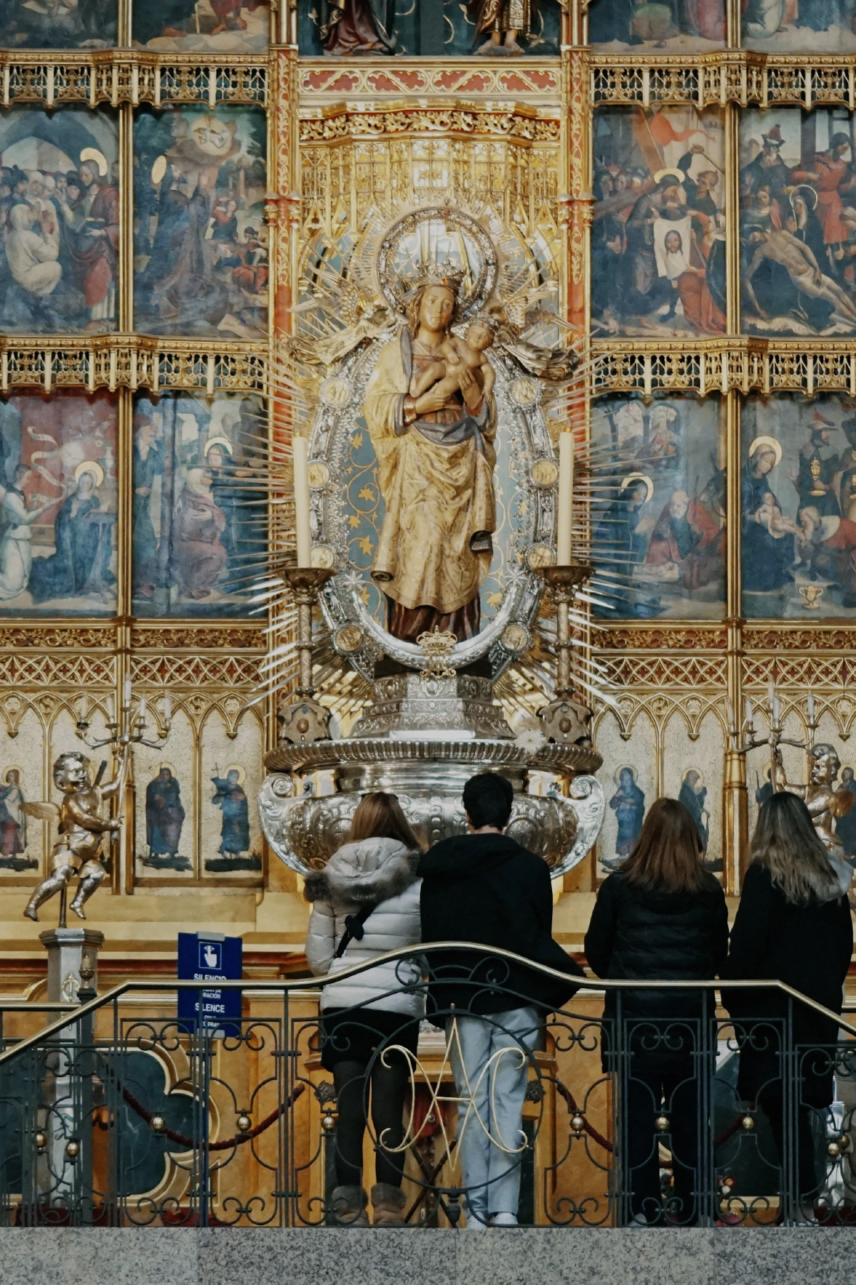 A gilded altar featuring a statue of the Virgin Mary holding the infant Jesus, with intricate religious artwork in the background and visitors observing.