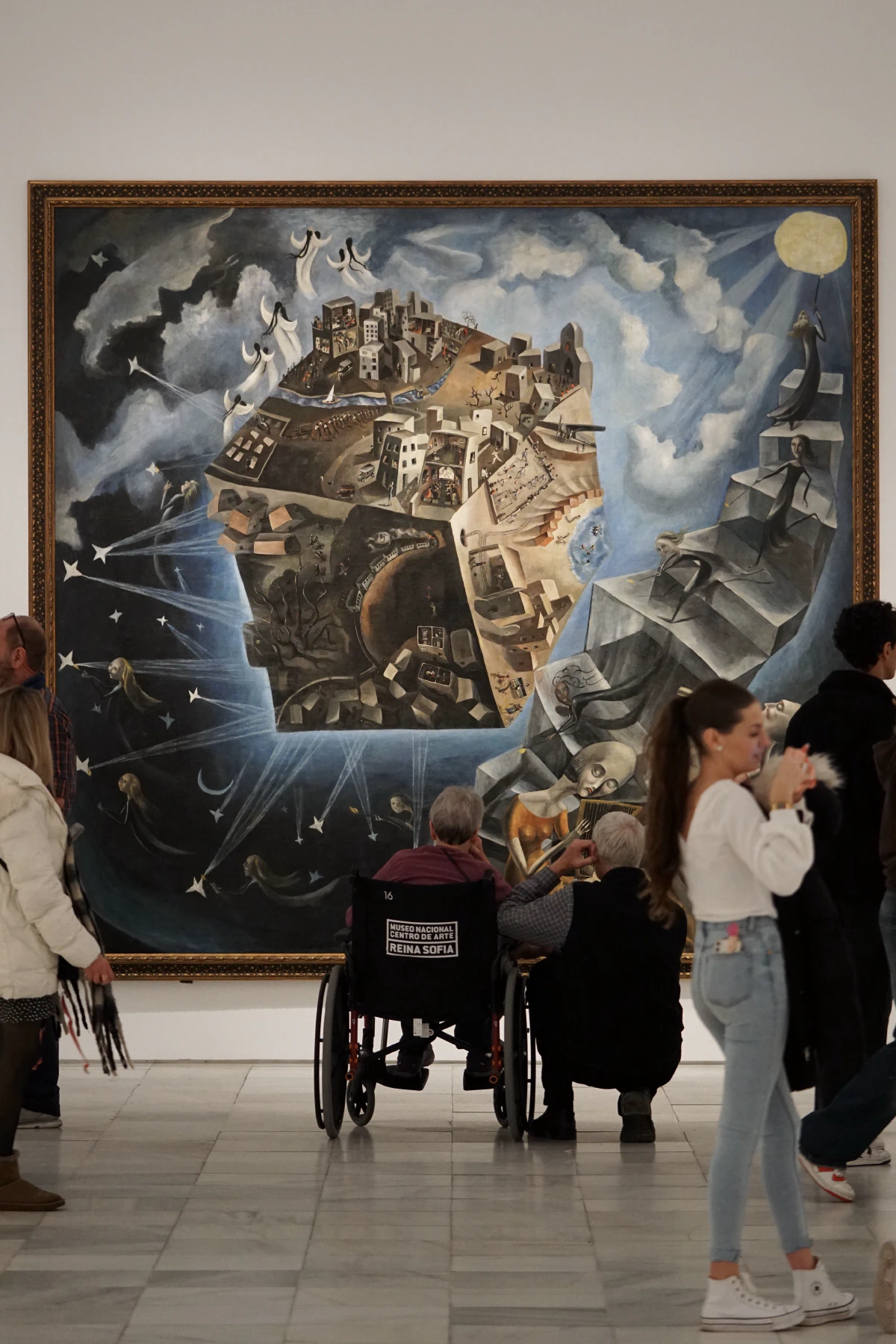 A surrealist painting of a floating cityscape with celestial imagery, displayed at the Reina Sofía Museum, observed by visitors including someone in a wheelchair.