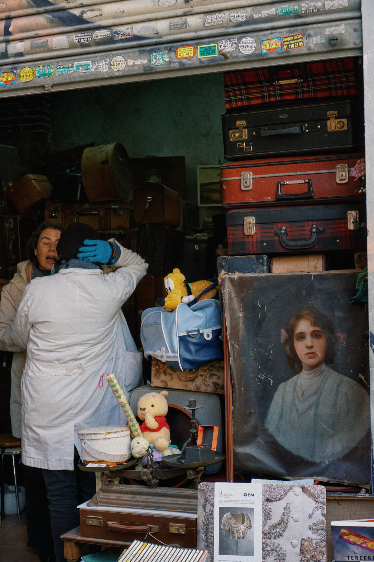 A vintage market stall with a collection of old suitcases, a painted portrait, stuffed toys, and various trinkets.