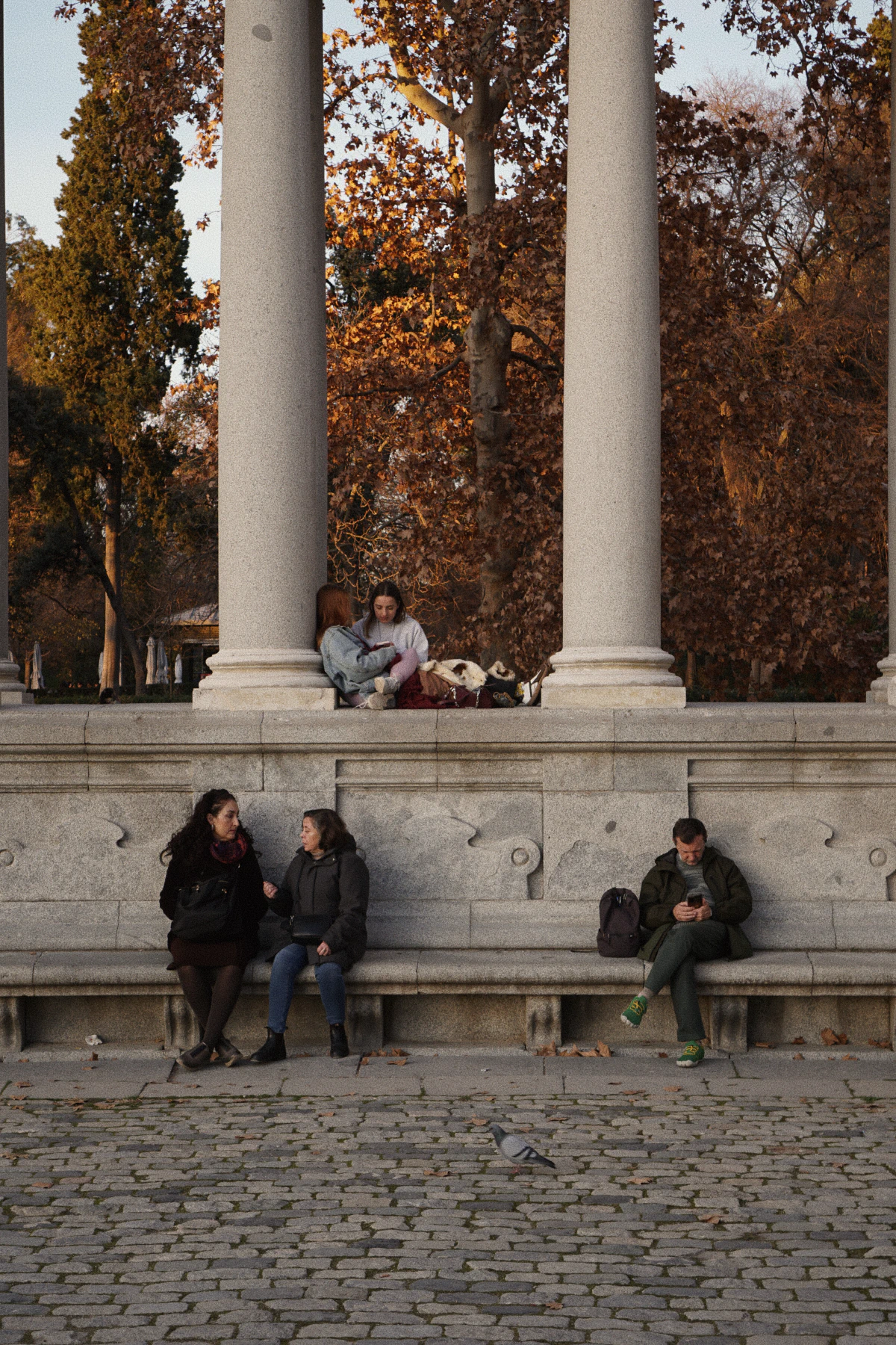 People sitting and chatting on a stone bench beneath tall columns in a park, framed by golden autumn trees.