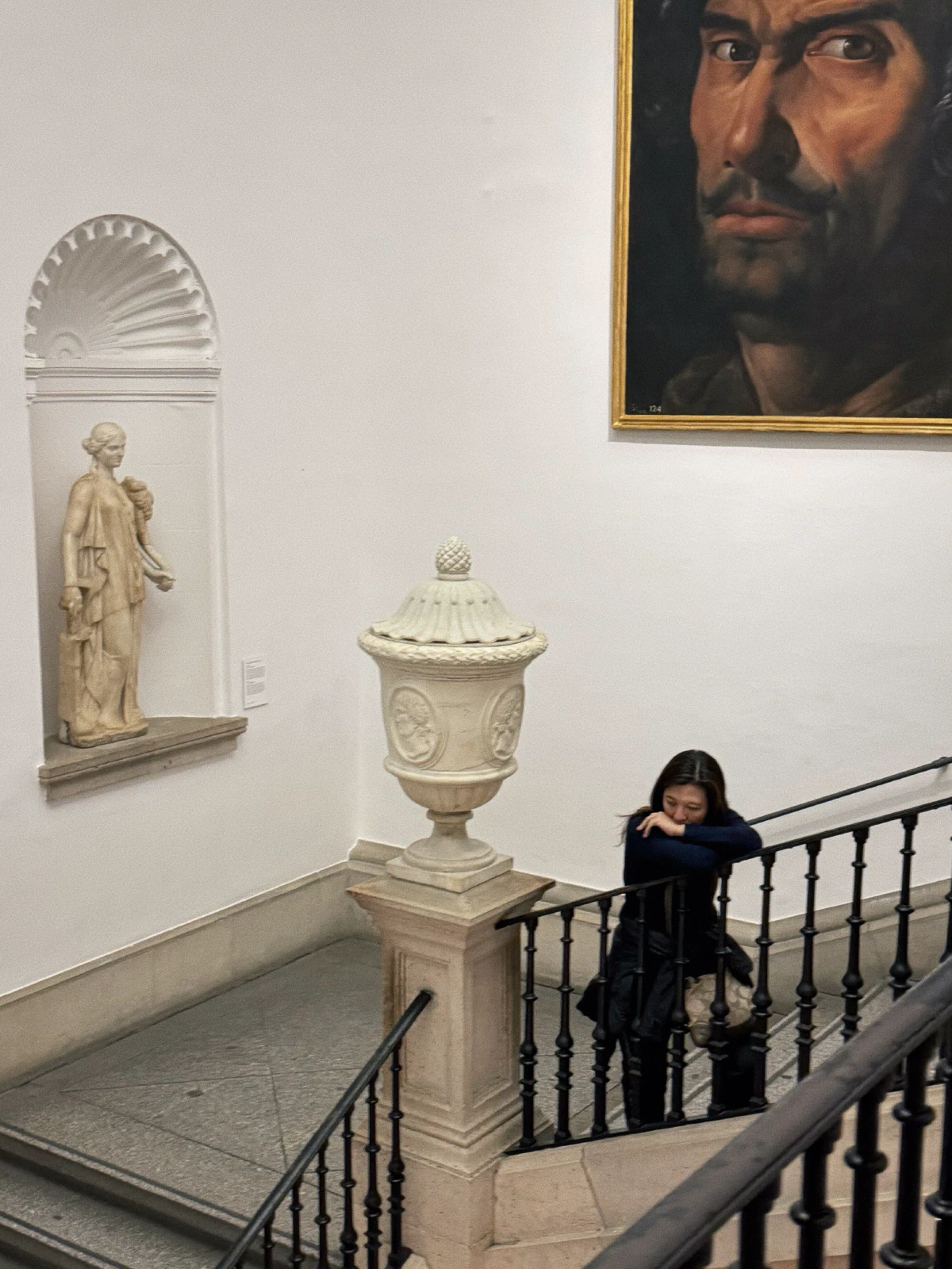 A thoughtful woman leaning on a staircase railing inside a museum, surrounded by classical art and sculptures.