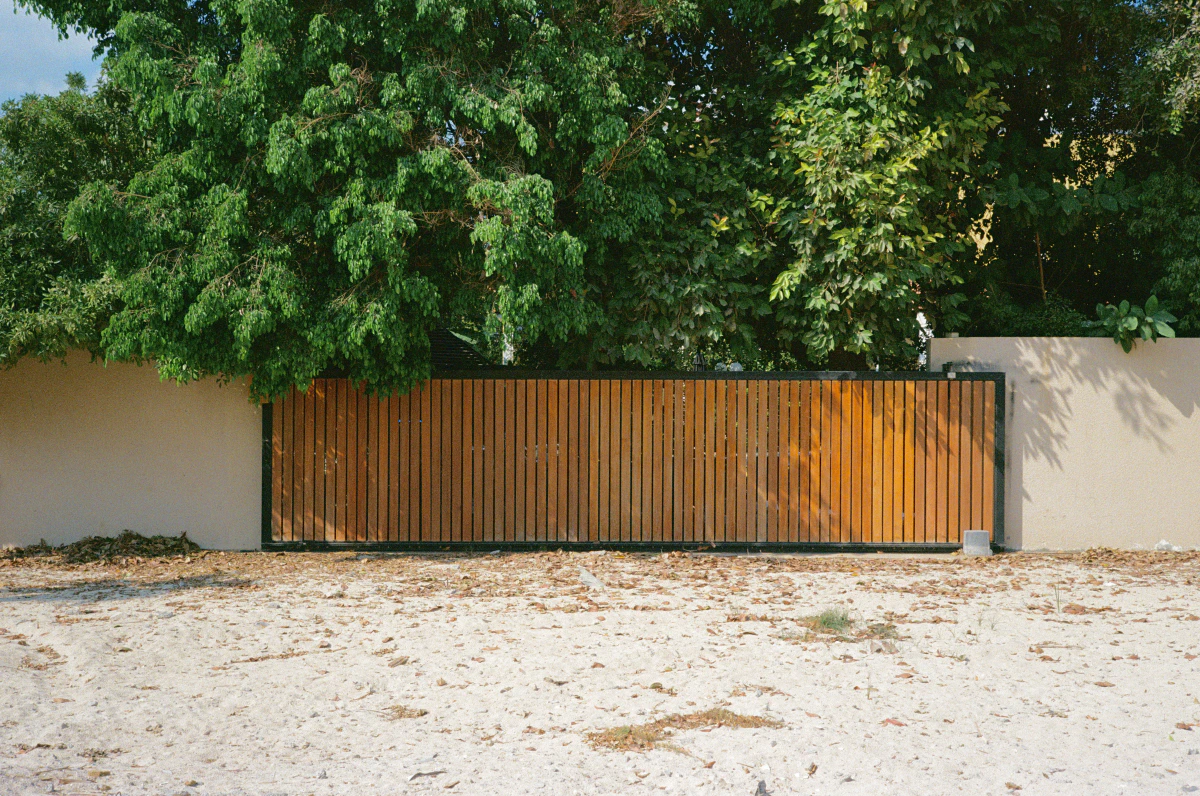 A tall wooden gate framed by lush green trees and beige walls, with a sandy ground in the foreground.