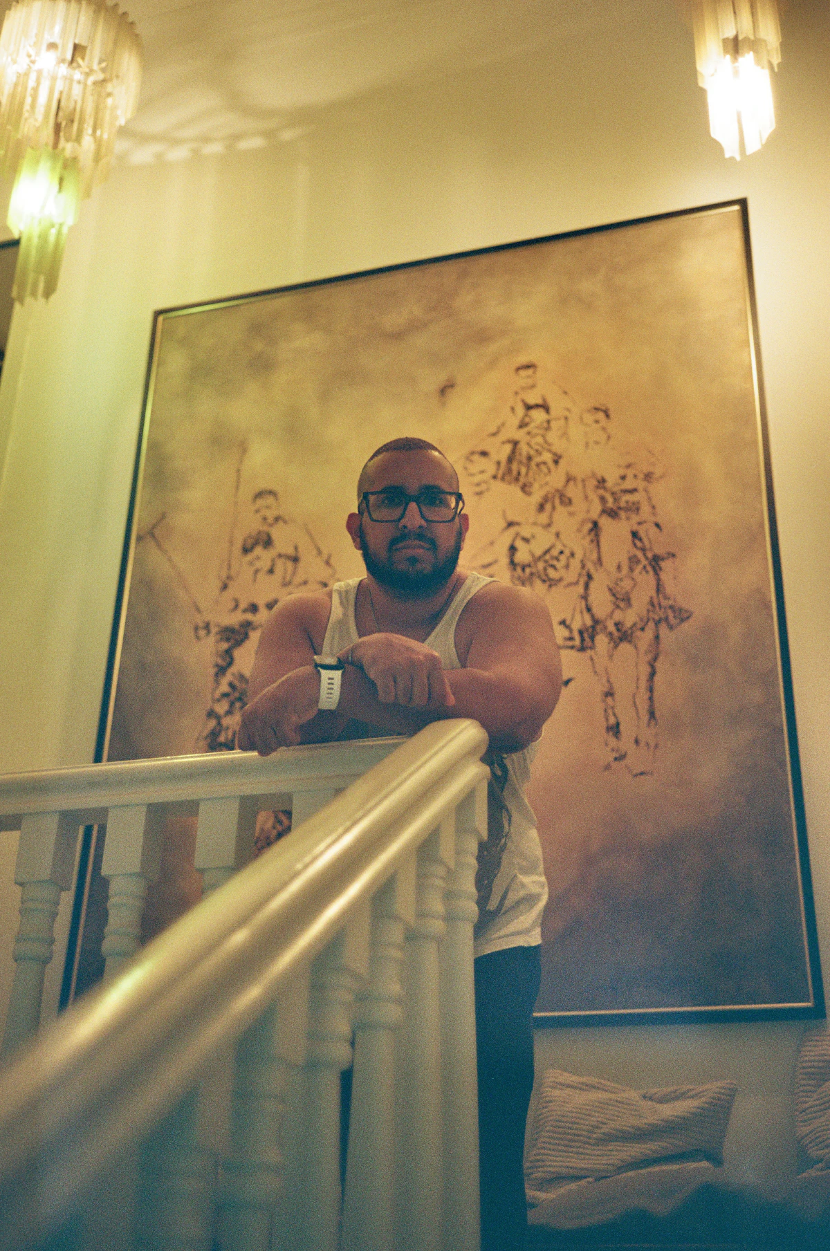 A man with glasses and a sleeveless shirt leaning on a staircase railing, with a large painting of soldiers on horseback behind him, illuminated by warm lights.