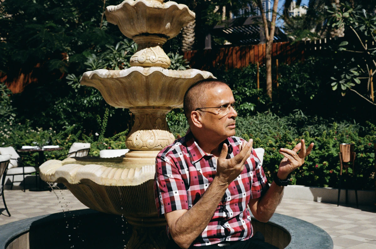 A man in a red and black checkered shirt gesturing while seated in front of a tiered stone fountain, surrounded by greenery on a sunny day.
