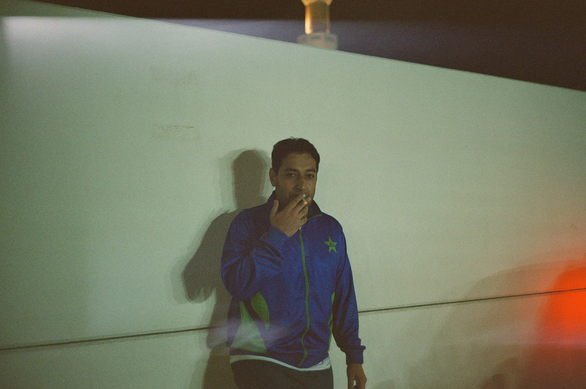 A man smoking a cigarette, wearing a blue jacket with the Pakistan national cricket team emblem, standing against a white wall at night, with faint red and yellow light reflections in the foreground.