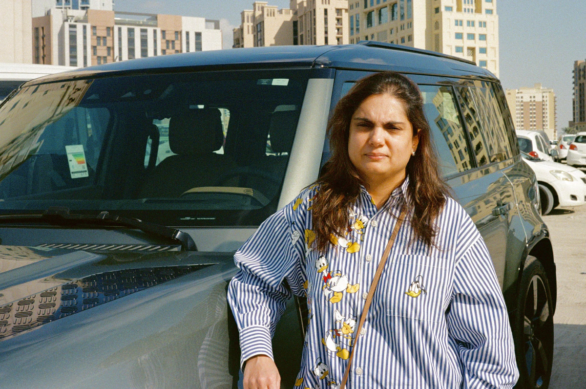 A woman in a striped shirt with cartoon characters standing next to a dark gray SUV in a sandy, sunlit parking lot, with buildings in the background.