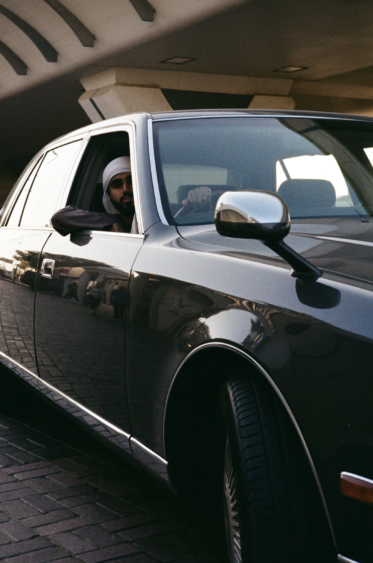 A man wearing traditional Middle Eastern attire, sitting inside a black Toyota Century with his hand on the steering wheel, reflecting the surroundings.