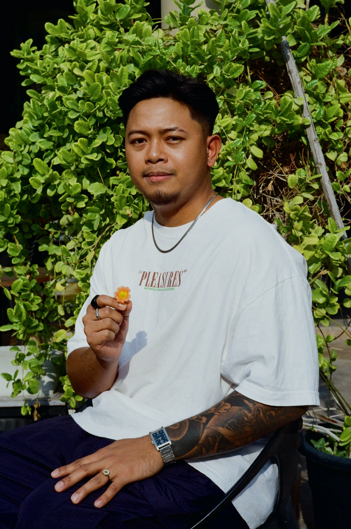 A man sitting in front of lush green plants, holding a small yellow flower, wearing a white shirt with ‘Pleasures’ written on it.