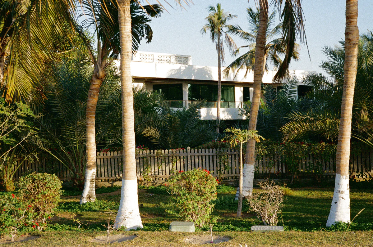 A villa surrounded by lush palm trees, a wooden picket fence, and neatly trimmed bushes under a sunny sky.