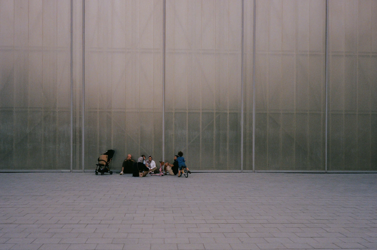 A small group of people, including children and adults, sit and play against the vast backdrop of a translucent, industrial-style wall. The open, empty plaza surrounding them emphasizes a sense of scale and space.