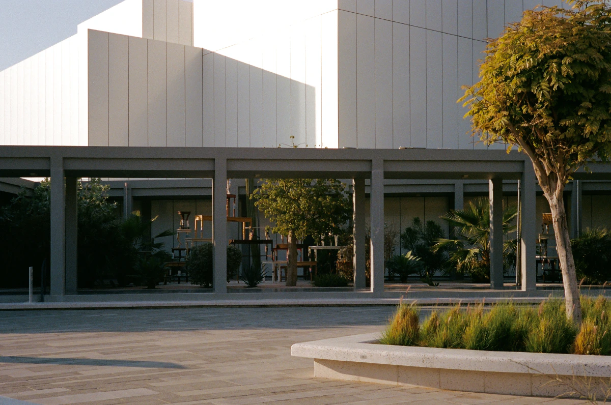 A modern architectural space bathed in warm sunlight, featuring a minimalist white building with clean geometric lines. A shaded walkway in the foreground frames lush greenery and sculptures, blending nature with contemporary design.