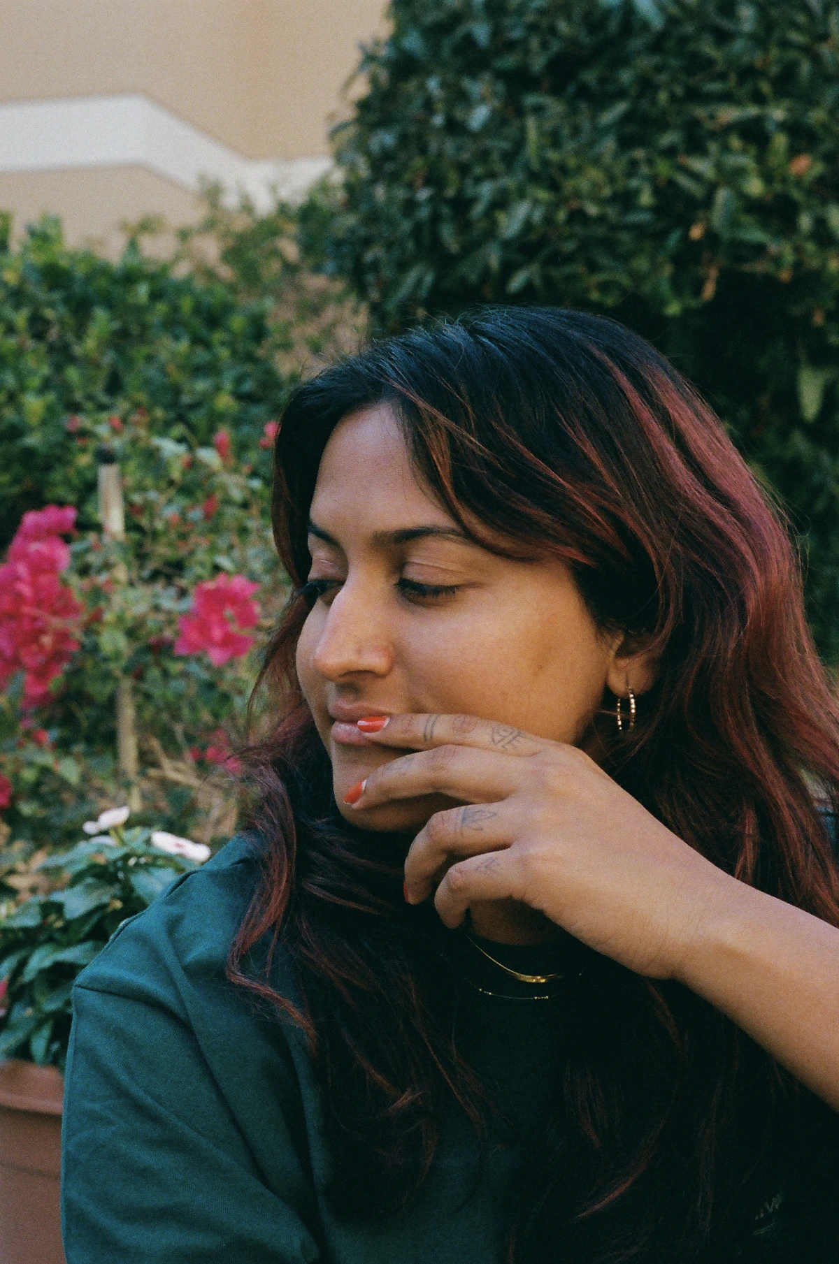 A woman with dark red-streaked hair sits in a garden, her face turned to the side in quiet thought. Her fingers rest gently against her lips, nails painted orange, with delicate tattoos on her hand.