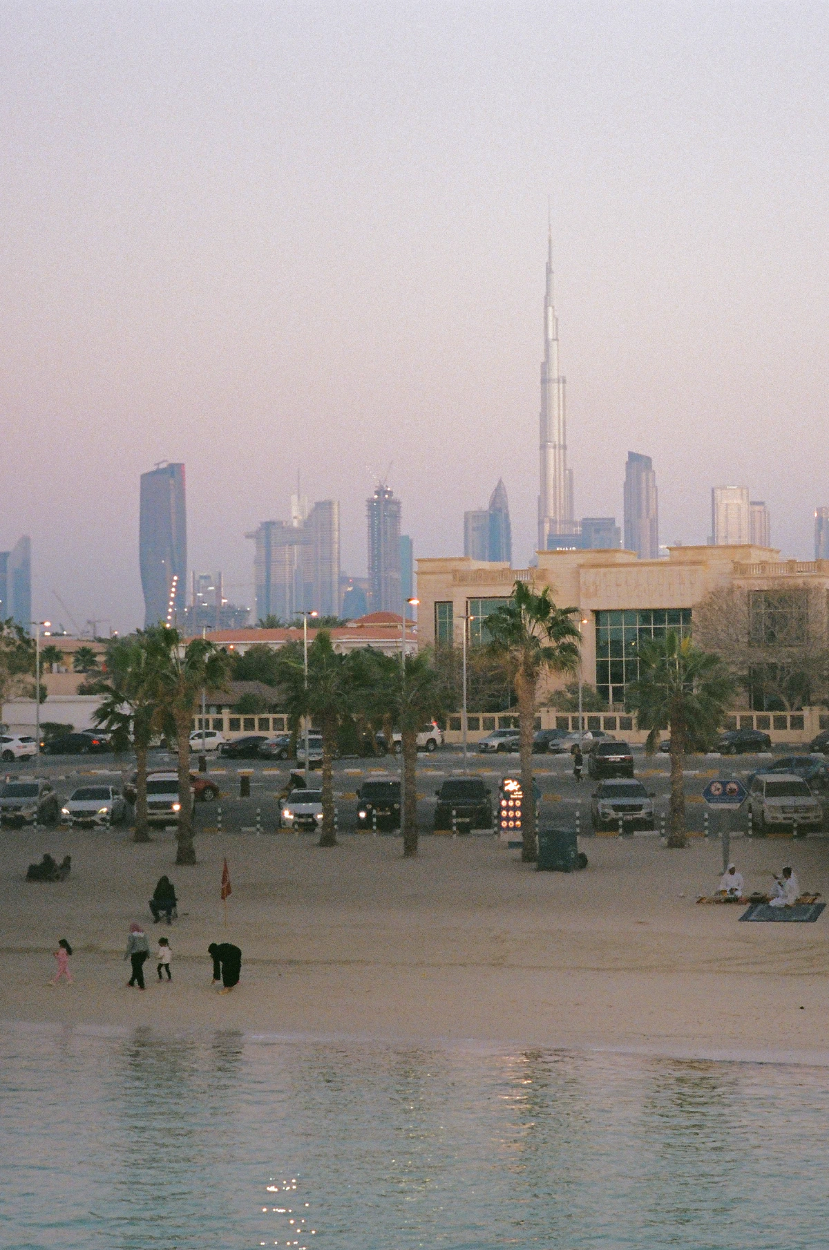 Serene beach scene at dusk with people strolling along the sand and sitting by the water. In the background, palm trees and parked cars line the shore, while the Dubai skyline, dominated by the towering Burj Khalifa, rises against a soft pastel sky.