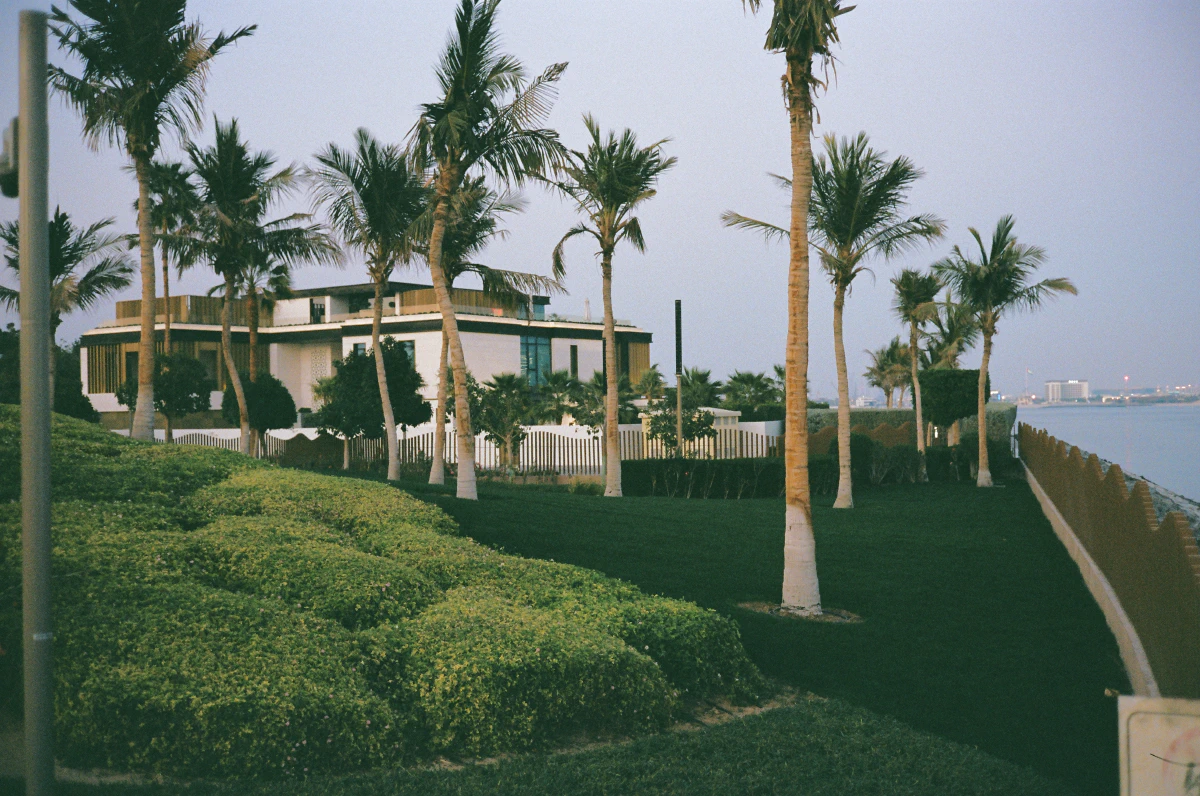 A modern white villa with wooden accents sits along a waterfront, surrounded by lush greenery and tall palm trees. A manicured lawn slopes toward a decorative fence that separates the property from the calm sea, with distant city lights glowing on the horizon.
