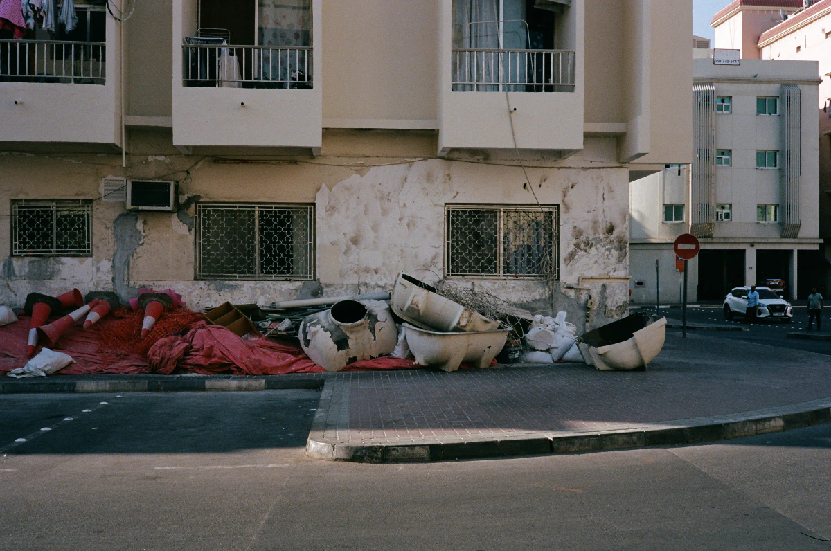 A worn-out residential building with peeling paint and exposed concrete stands above a pile of discarded construction debris, including broken planters, tangled wires, and traffic cones draped over a red tarp. In the background, a quiet street with parked cars and pedestrians contrasts with the chaotic scene below.