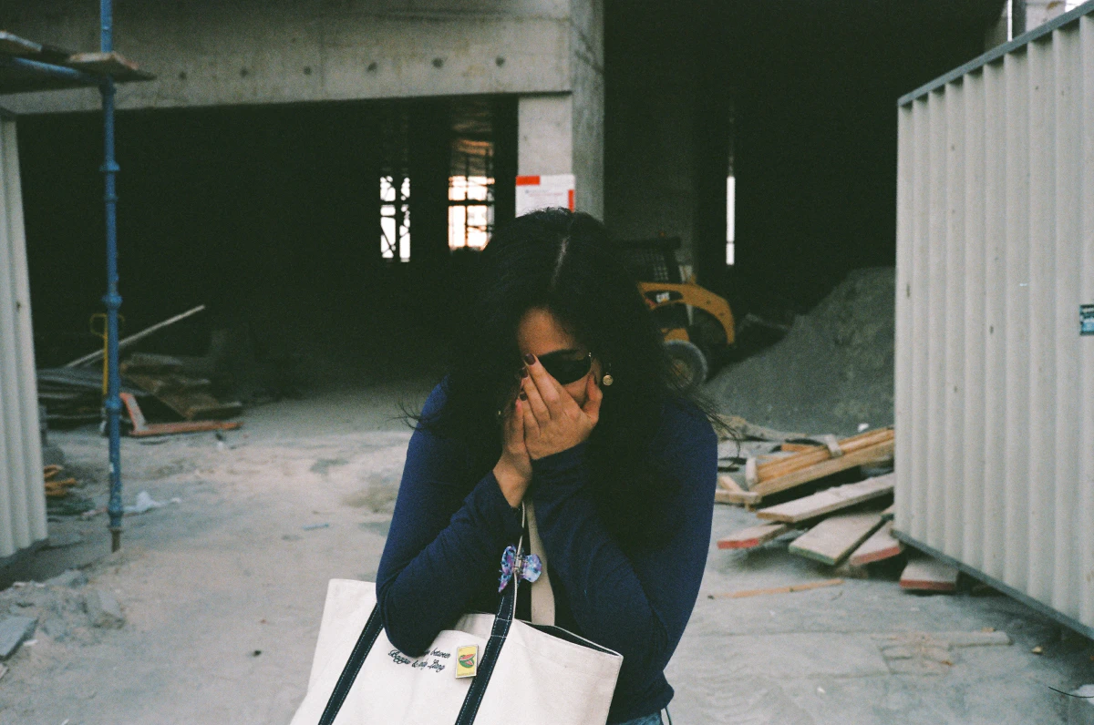 A woman in a deep blue top stands in front of a dimly lit construction site, covering her face with both hands. She carries a white tote bag adorned with a small watermelon pin, a symbol of solidarity with Palestine. Behind her, scaffolding, wooden planks, and a parked loader frame the unfinished space.