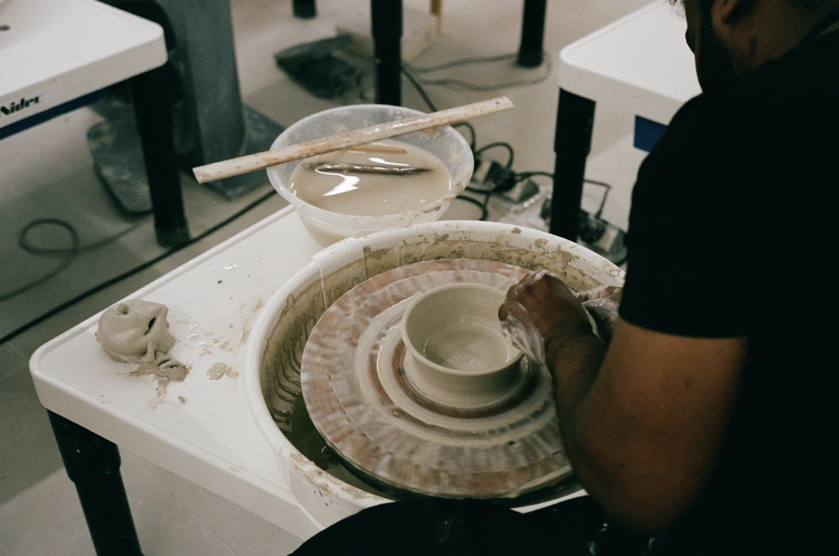 A potter’s hands shape a spinning clay vessel on a pottery wheel, the wet clay glistening under soft lighting. Nearby, a bowl of water and a wooden tool rest on a white table, hinting at the careful process of creation.