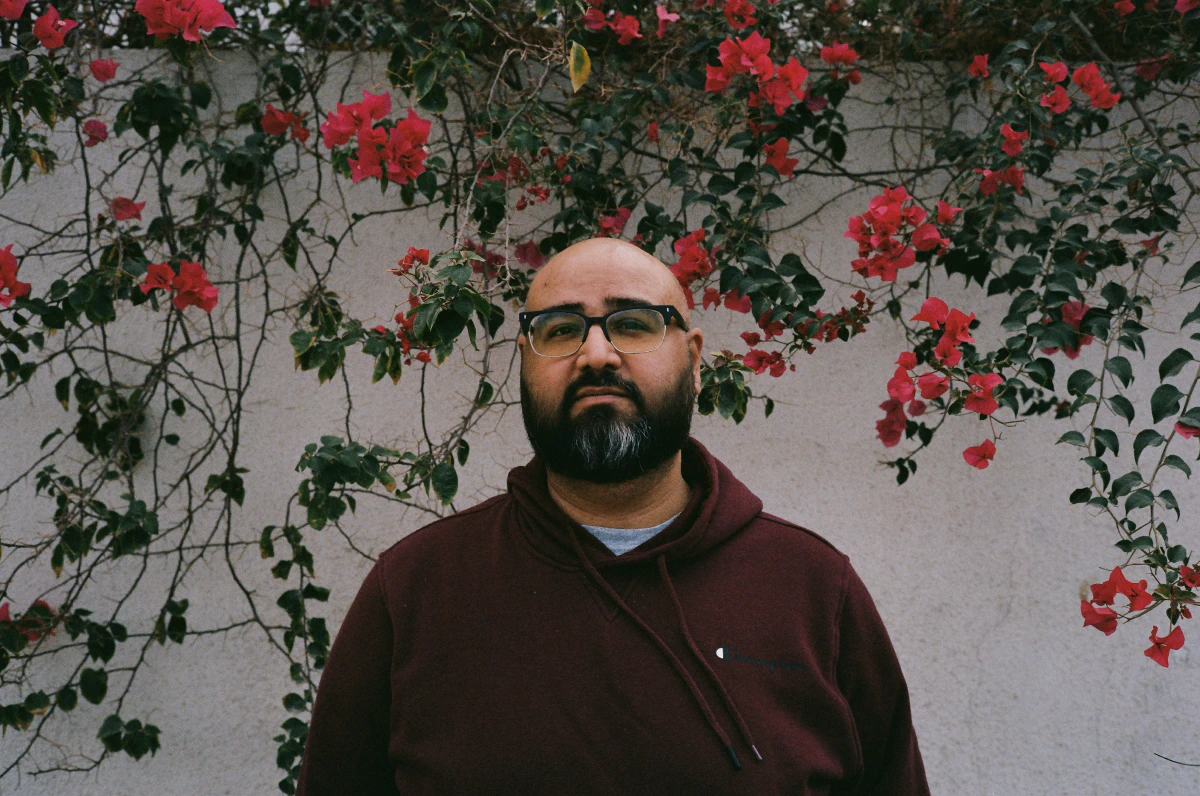 A bearded man in a maroon hoodie stands against a white wall covered in climbing bougainvillea, its vivid red flowers framing him. He gazes at the camera with a calm yet introspective expression.