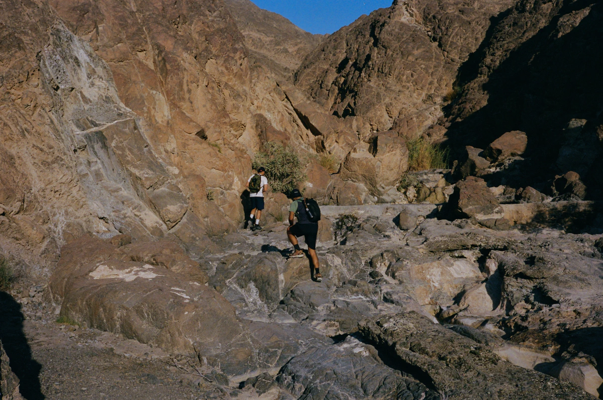Two hikers navigate the rocky terrain of the Al Dahir trail, stepping carefully along uneven stone surfaces. The mountains rise steeply around them, with sunlight casting deep shadows across the rugged path.