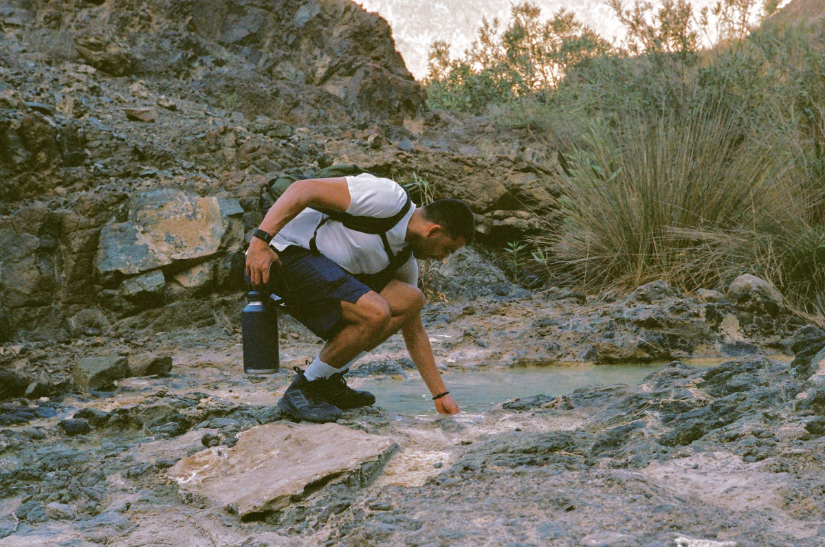A hiker crouches down to touch the surface of a small water pool nestled among the rocks. Wearing a white t-shirt and carrying a backpack, he holds a water bottle in one hand while reaching out to feel the coolness of the stream.