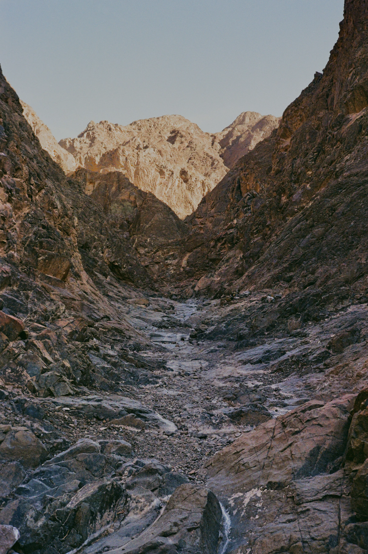 A narrow, dry wadi snakes between steep rock faces, with golden light catching the upper mountain ridges in the background.