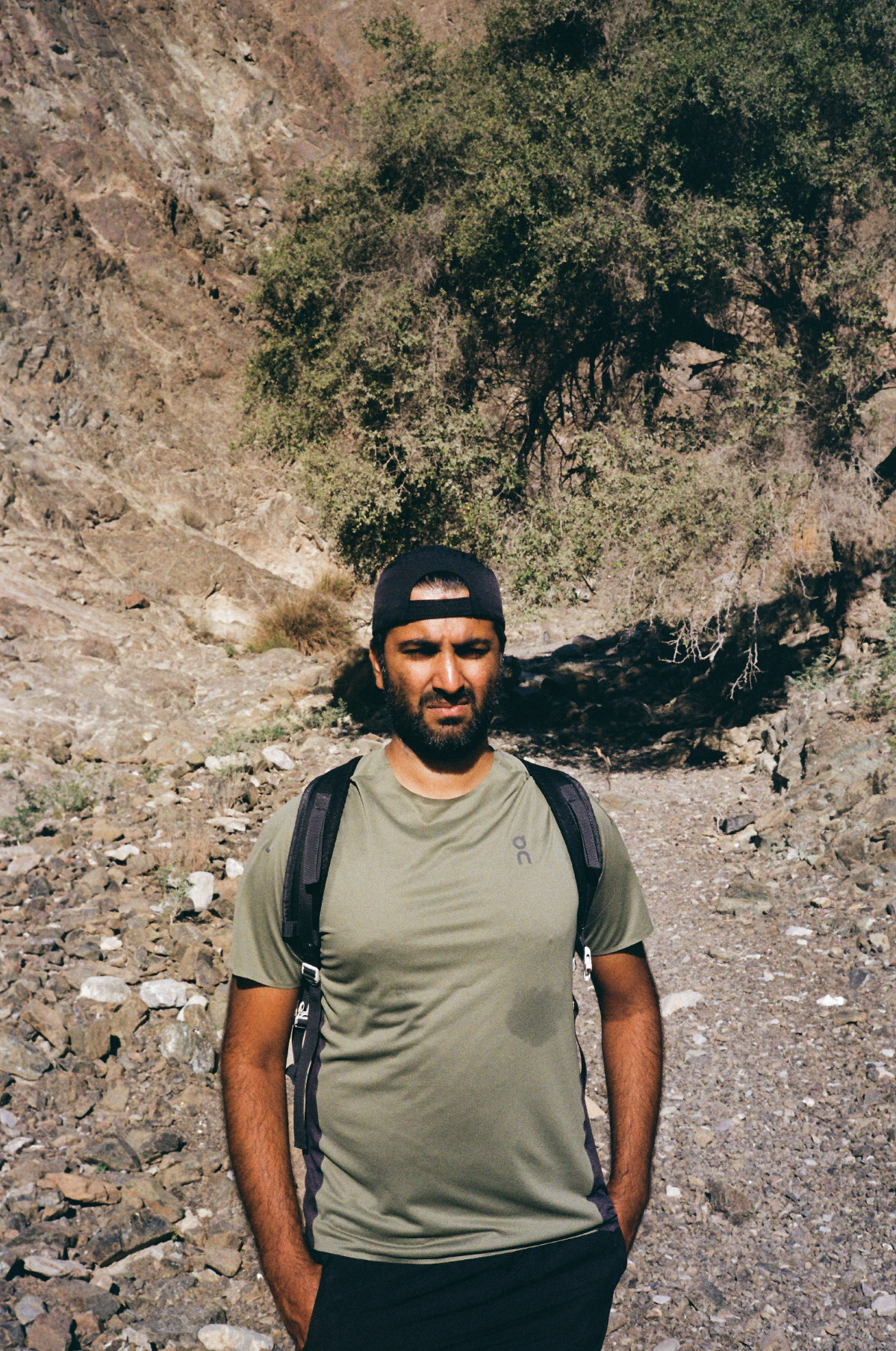 My cousin Nikhil standing on a rocky path with a shaded tree and rugged cliffside behind him.