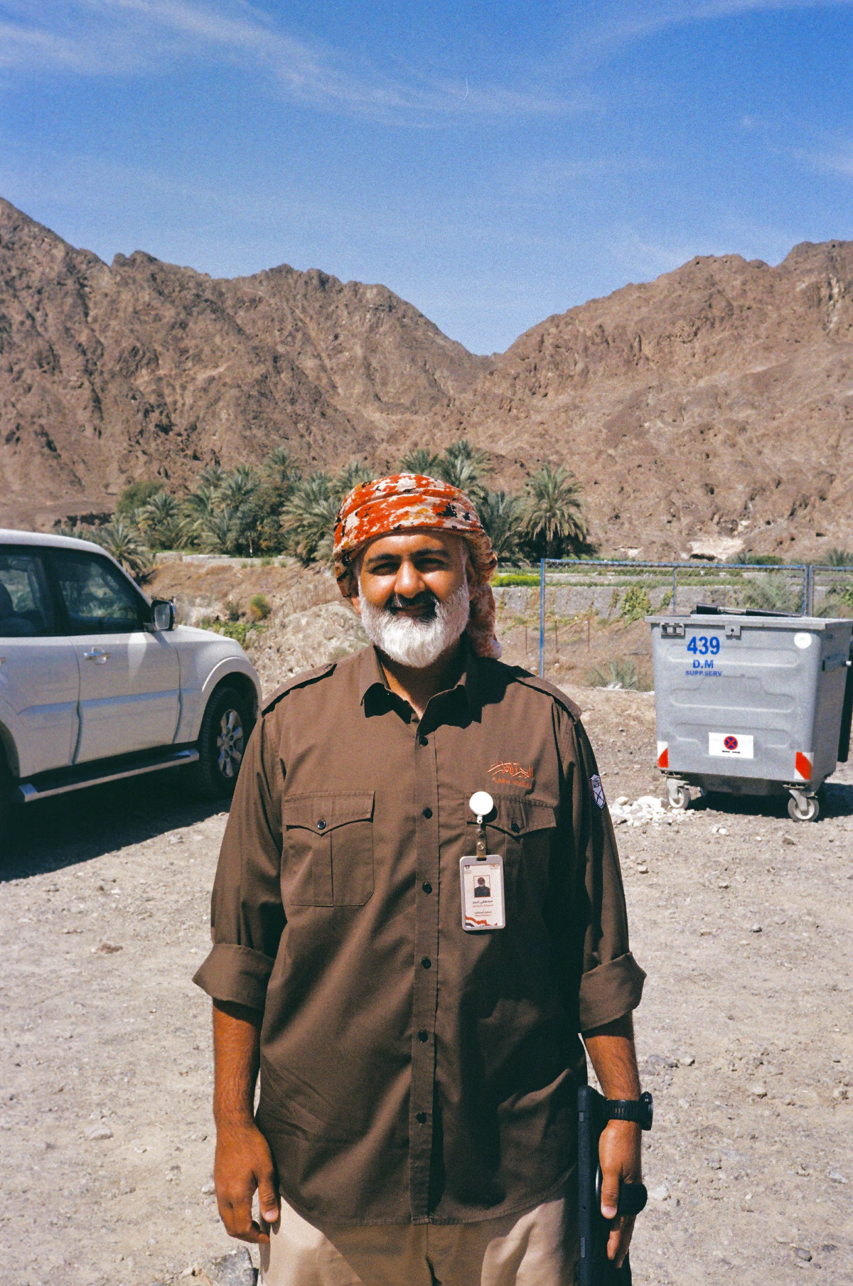Mustafa, from Fujairah Adventures, stands smiling in front of rocky desert mountains and palm trees, under a clear blue sky. We met him at the end of our hike and talked about our shared love of classic cameras.