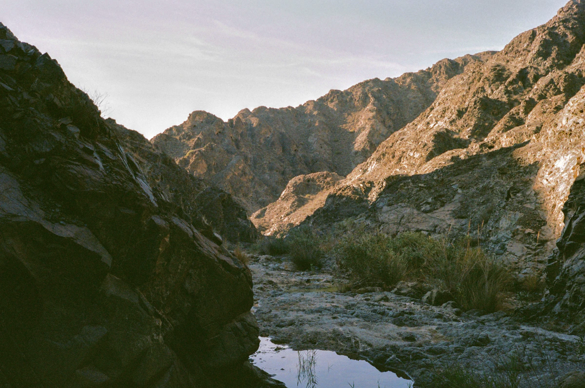 A rocky mountain landscape at the Al Dahir trail in Fujairah, UAE. Jagged peaks rise in the background, their surfaces illuminated by soft sunlight. A shallow stream reflects the sky, winding through the rugged terrain, with patches of dry grass and shrubs scattered along the path.